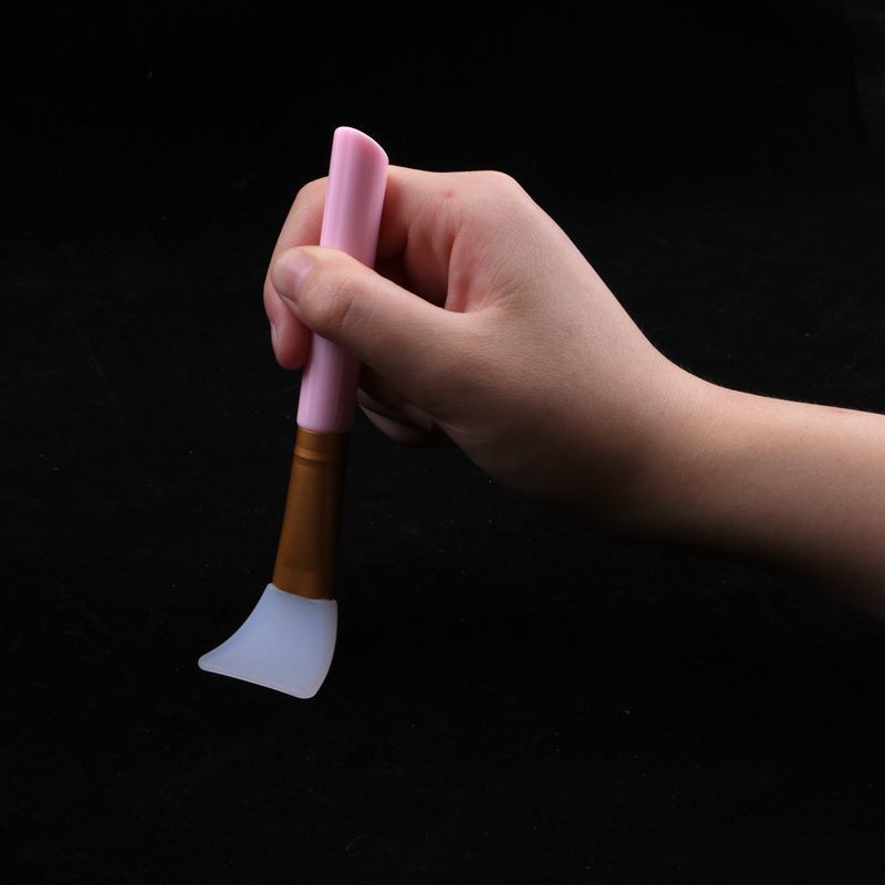 Hand holding a silicone facial mask brush with a pink handle, gold ferrule, and flexible white silicone head.