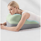 A woman rests chest-down on a contoured, two-toned mint green and grey foam support cushion on a white bed.