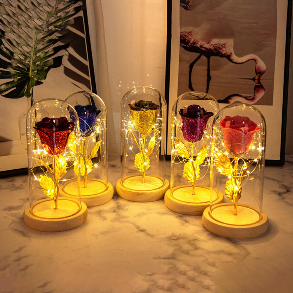 Five glittery roses in varied colors encased in glass domes on wooden bases, illuminated by warm LED fairy lights.
