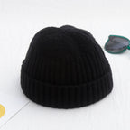 A black, ribbed, short-cuff knit beanie on a white surface, next to green-rimmed sunglasses.
