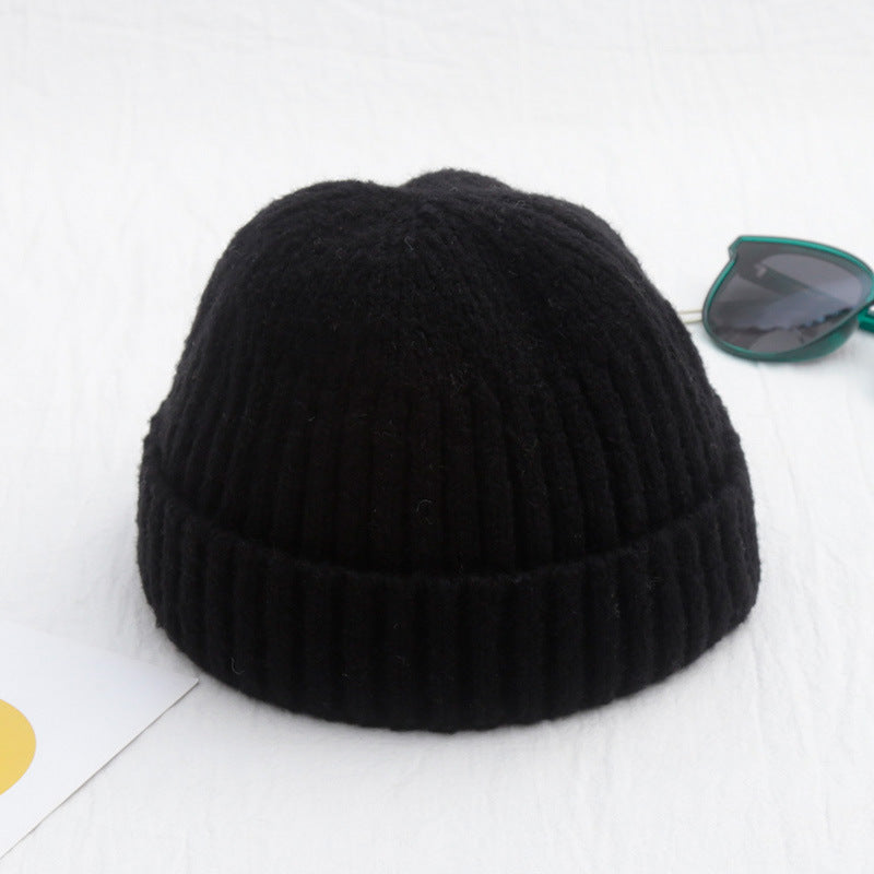 A black, ribbed, short-cuff knit beanie on a white surface, next to green-rimmed sunglasses.
