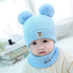 Baby wearing a light blue ribbed wool hat with two faux-fur pom-poms and a matching scarf with embroidered text.
