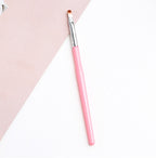 A makeup brush with a bright pink handle, silver ferrule, and soft brown synthetic bristles on a pink and white surface.