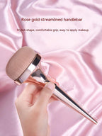 Hand holding a rose gold makeup brush with a dense, rounded head against a soft pink satin background.