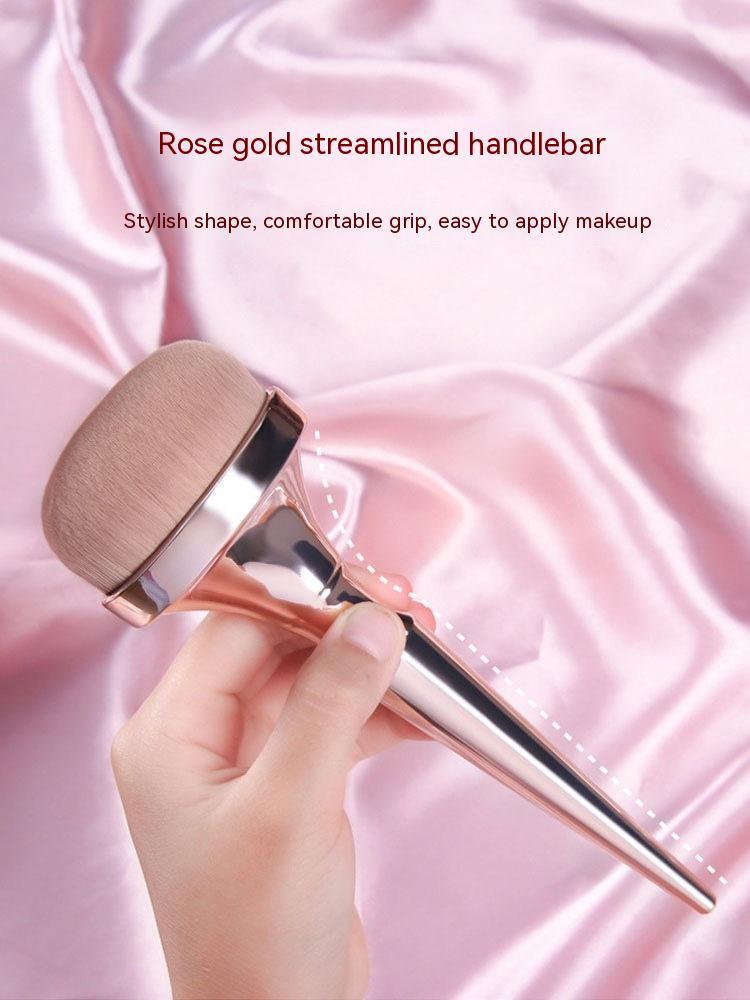 Hand holding a rose gold makeup brush with a dense, rounded head against a soft pink satin background.