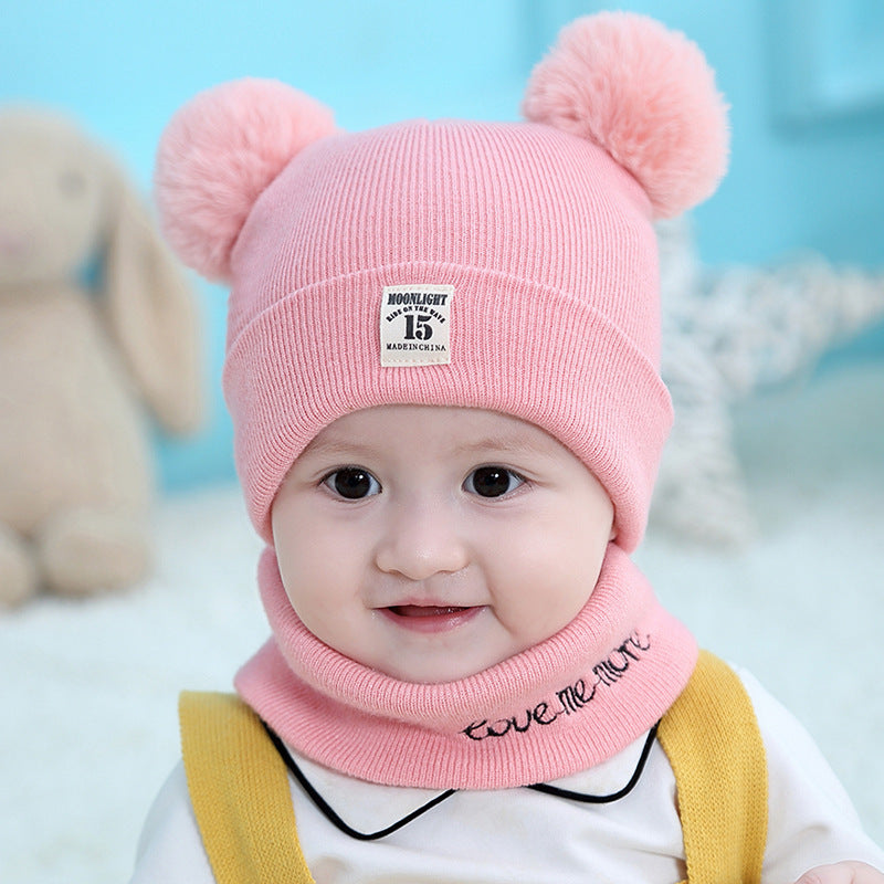 A baby wears a pink ribbed wool hat with two plush pom-poms and a matching scarf with "love me more" embroidery.