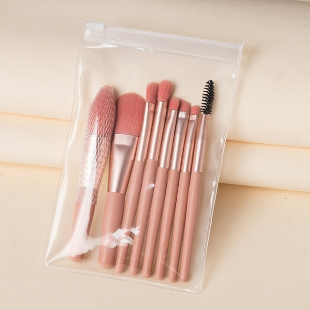 Set of 8 mini pink makeup brushes with gold ferrules in a clear, zippered travel pouch against a beige background.