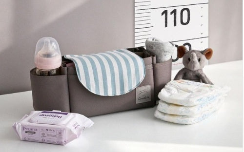 Grey fabric stroller organizer with blue-striped flap, holding baby bottle, diapers, and toy on a white surface.