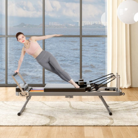 Woman doing pilates on a sleek, black foldable reformer machine with a metal frame in a bright room overlooking water.