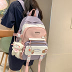 A Korean-style, pastel pink and purple fabric backpack with cartoon pins and a fluffy bear charm hanging from the side.