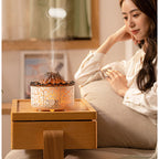 A woman sits by a glowing, white crackled volcano-shaped humidifier emitting a jellyfish-like steam ring on a wood tray.