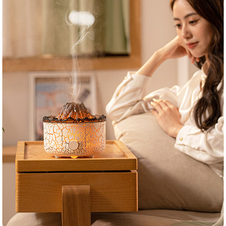 A woman sits by a glowing, white crackled volcano-shaped humidifier emitting a jellyfish-like steam ring on a wood tray.