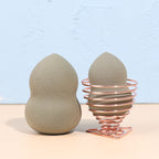 Two taupe makeup sponges, one resting in a rose gold coiled wire stand on a beige surface against a blue background.