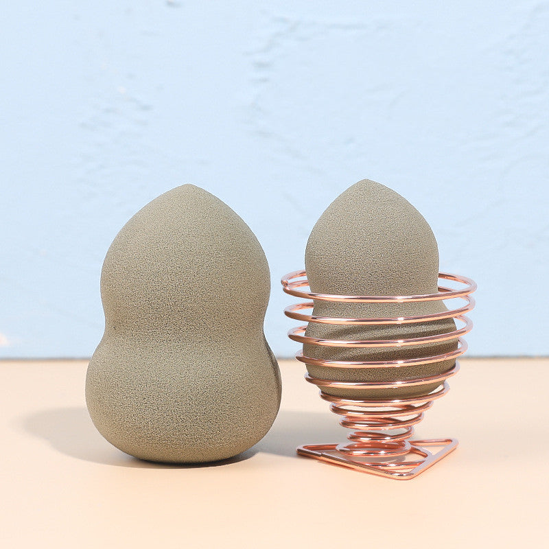 Two taupe makeup sponges, one resting in a rose gold coiled wire stand on a beige surface against a blue background.