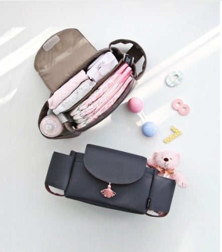 Two gray fabric stroller organizer bags, one open showing baby supplies and one closed with a tassel, near baby toys.