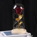 A glittering red rose with gold leaves and LED fairy lights inside a glass dome on a wooden base.