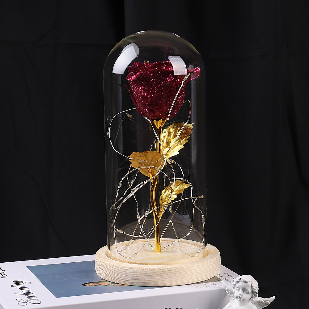 A glittering red rose with gold leaves and LED fairy lights inside a glass dome on a wooden base.