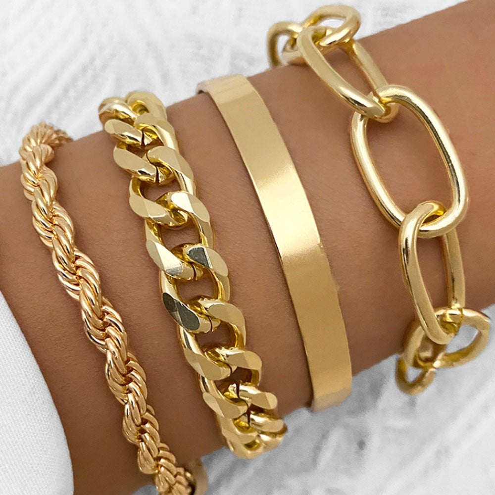 A set of four gold-toned hip hop bracelets on a wrist, featuring rope, cuban link, smooth cuff, and oval link chains.