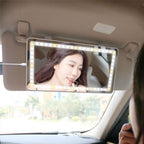 White rectangular car visor mirror with bright LED border, attached to a beige car visor, reflecting a woman applying makeup.
