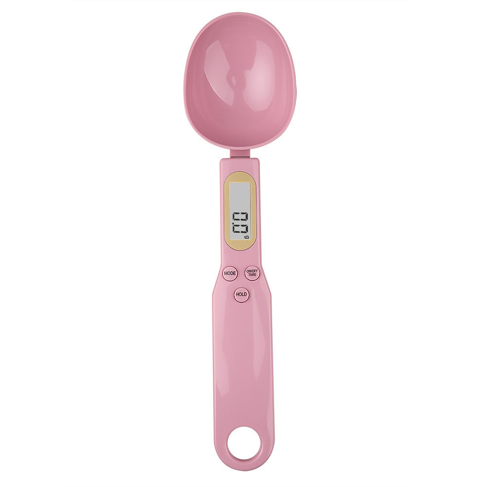 Pink plastic digital spoon scale with an LCD screen, measurement buttons, and a hanging hole at the base.