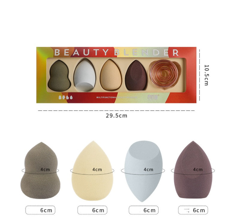 Four makeup sponges in various shapes and earthy colors, plus a metal coil stand in a labeled retail display box.