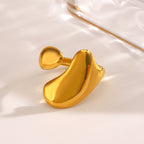 Gold-tone stainless steel ring with an open, asymmetric, organic liquid-shaped design on a reflective surface.