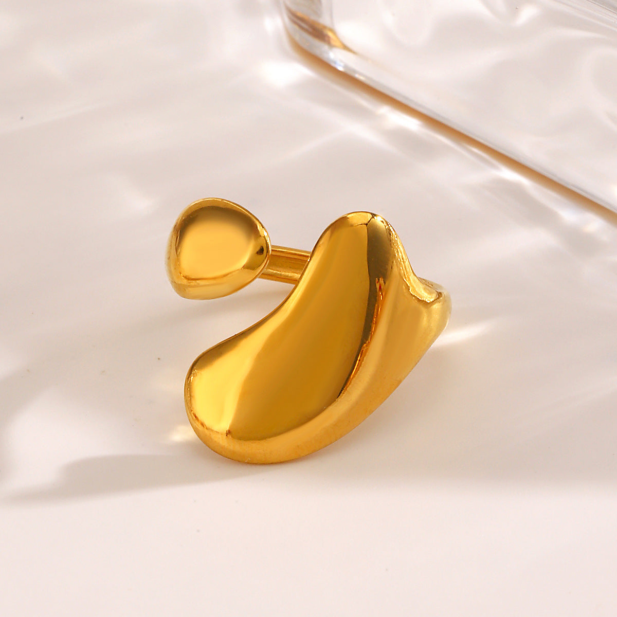 Gold-tone stainless steel ring with an open, asymmetric, organic liquid-shaped design on a reflective surface.
