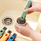A hand uses a green 5-in-1 multi-functional cleaning brush to scrub the rim of a small cup over a kitchen sink.