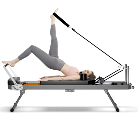 A person uses a foldable black Pilates reformer with metal frames and pulleys to perform a leg exercise.