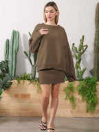 Woman wearing an olive green knit sweater and matching mini skirt, posing in front of cacti and greenery.