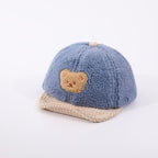 Blue fuzzy fleece baby hat with a cute teddy bear patch and a beige corduroy brim on a white background.