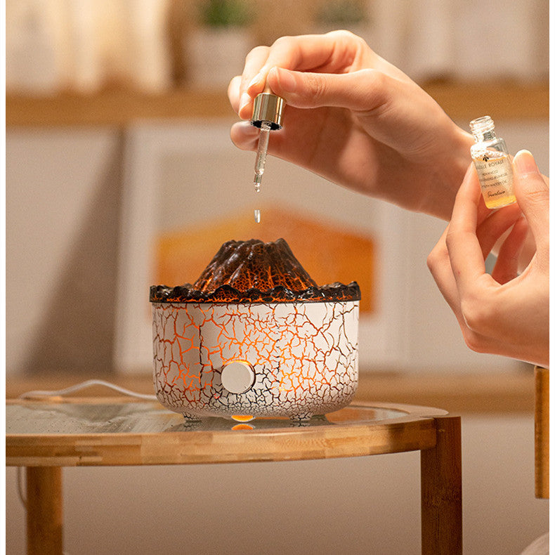 Volcano-shaped aroma diffuser with white crackle texture, glowing orange light, and a hand adding oil via dropper.