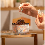 Volcano-shaped aroma diffuser with white crackle texture, glowing orange light, and a hand adding oil via dropper.