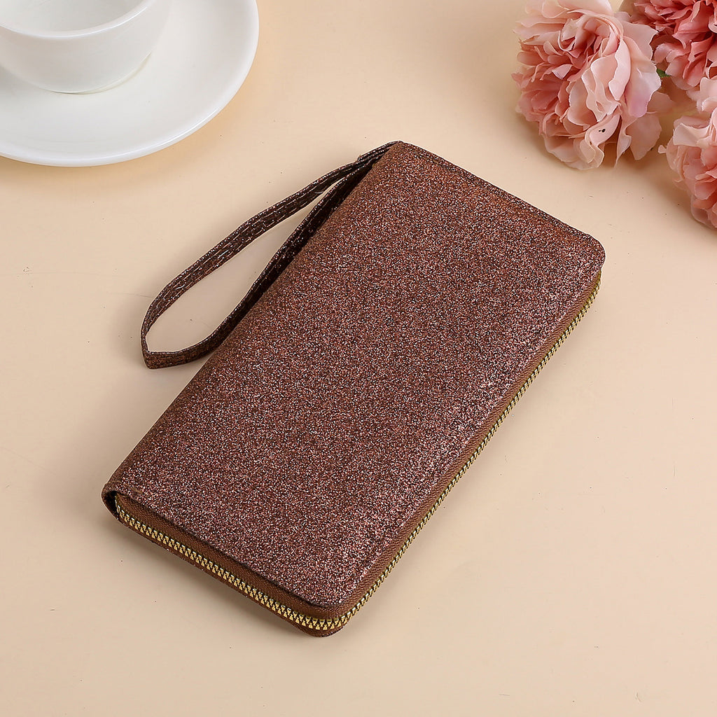 A brown, glittery rectangular clutch with a wrist strap and gold zipper, set on a surface with a coffee cup and flowers.