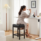 Black saddle bar stool with a sturdy steel frame and tufted seat, used by a woman applying makeup at a desk.