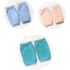 Soft terry fabric baby knee pads in light blue, peach, and teal, each featuring a non-slip rubber smiley face grip.