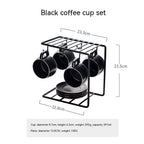 A matte black ceramic coffee cup set with gold rims, hanging on a matching black wire rack with a stack of saucers.