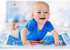 Smiling baby in blue onesie on a clear, inflatable water play mat with colorful, floating sea creature toys inside.