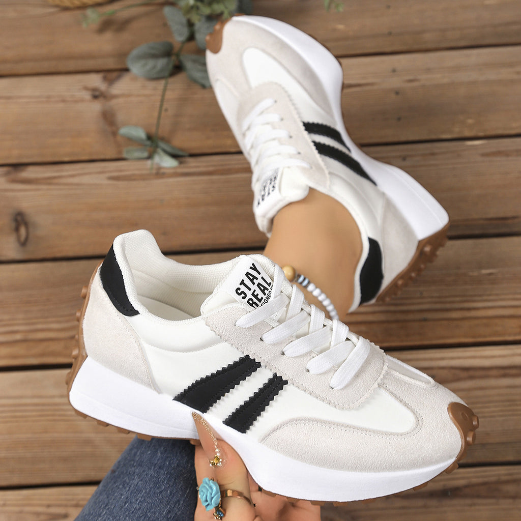 White and beige lace-up sneakers with black side stripes, thick white midsoles, and brown lug soles on a wood background.