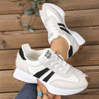 White and beige lace-up sneakers with black side stripes, thick white midsoles, and brown lug soles on a wood background.