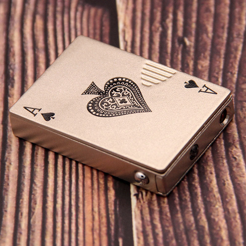 Matte gold metal lighter shaped like an Ace of Spades playing card with intricate engravings on a wood surface.