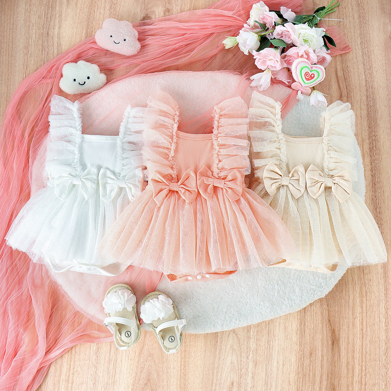Three baby jumpsuits in white, peach, and beige featuring tulle ruffles, bow accents, and snap-button bottoms.