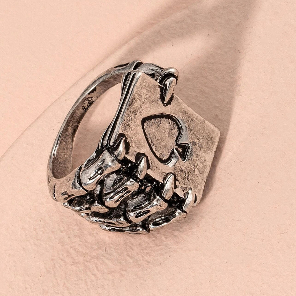 Silver-tone hip-hop ring featuring a skeleton hand clutching an ace of spades playing card on a soft pink background.