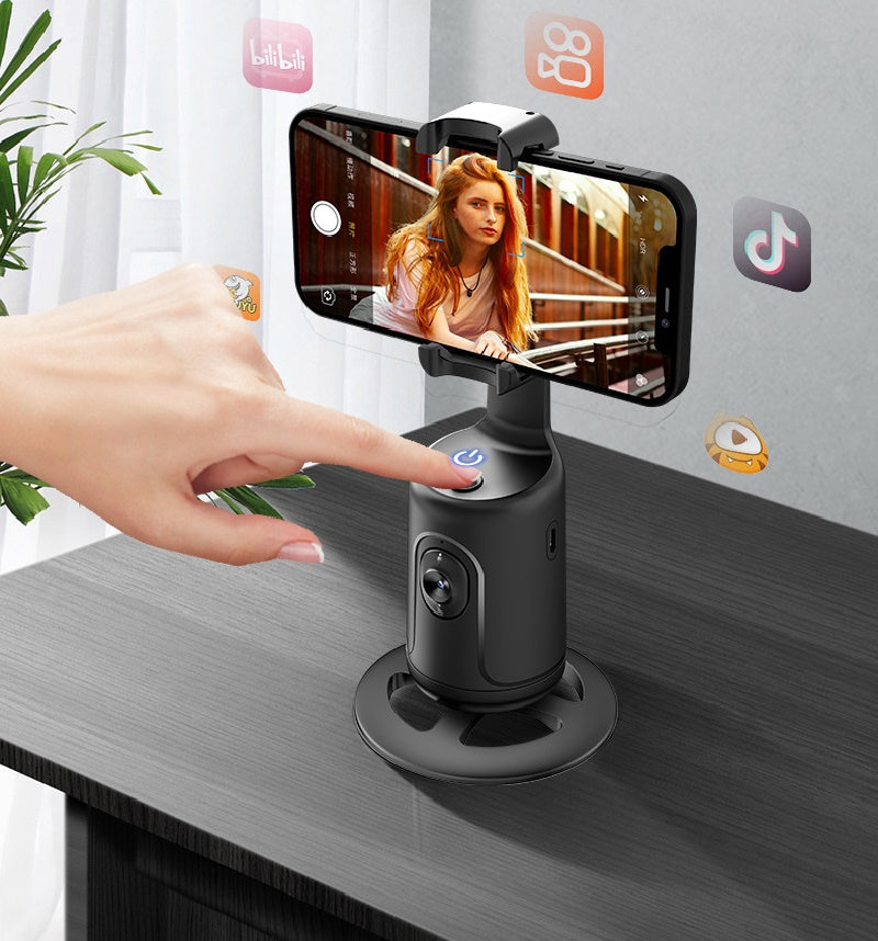 Black 360-degree auto-tracking phone gimbal on a desk with a hand pressing the power button and smartphone mounted.