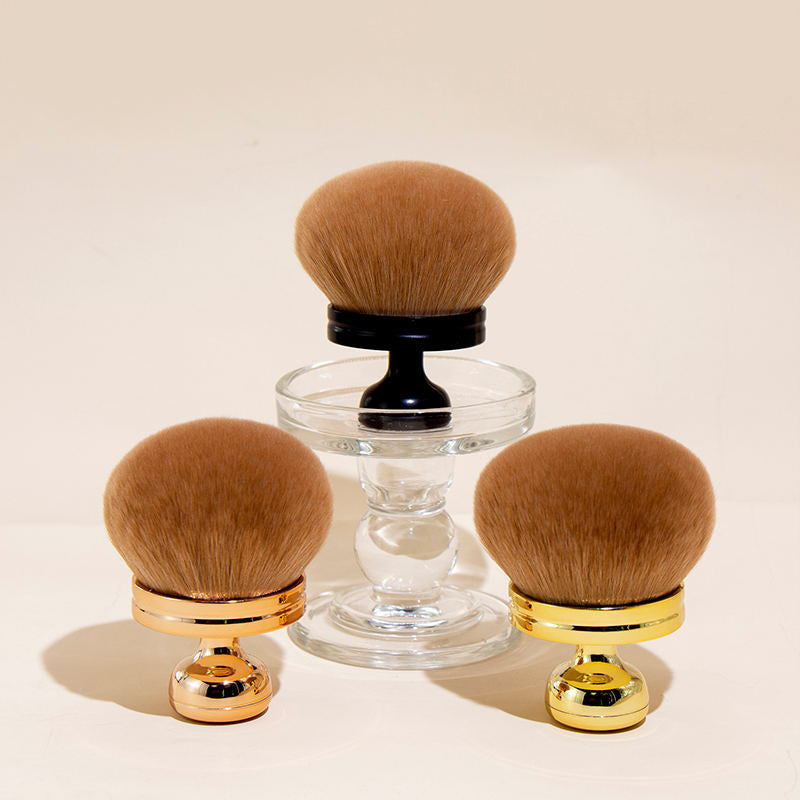 Three oversized, soft brown kabuki powder brushes with metallic gold, silver, and black handles on a glass stand.