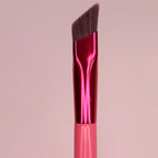 Angled eyebrow brush with dark brown synthetic bristles, shiny metallic red ferrule, and pink handle on a pink background.