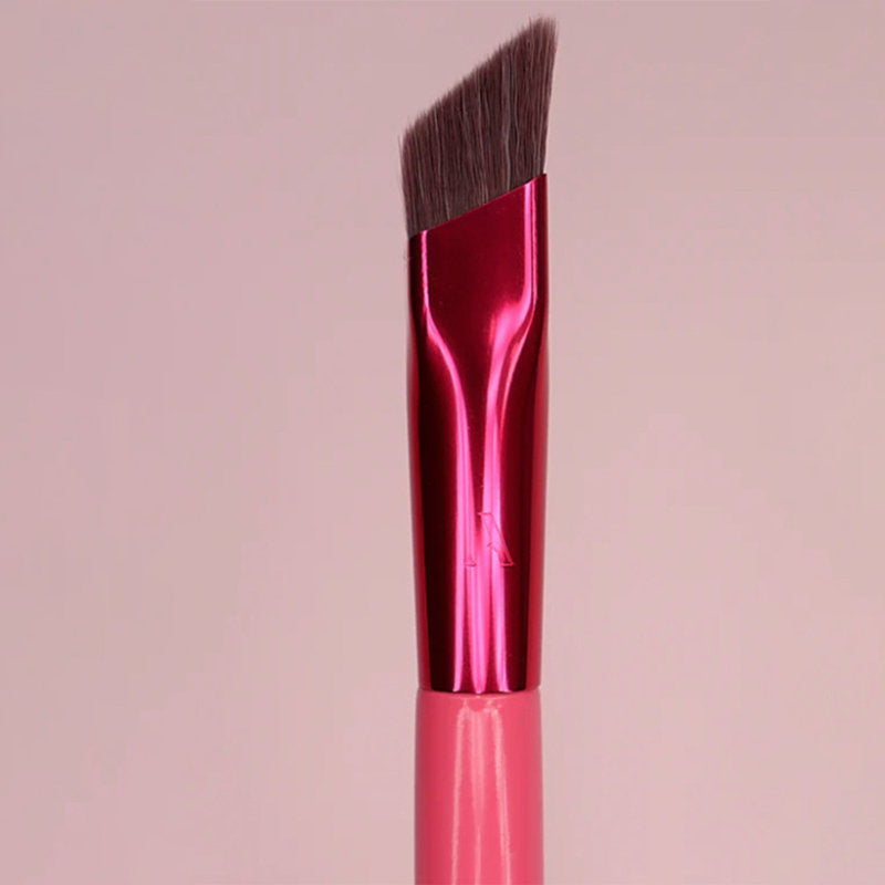 Angled eyebrow brush with dark brown synthetic bristles, shiny metallic red ferrule, and pink handle on a pink background.