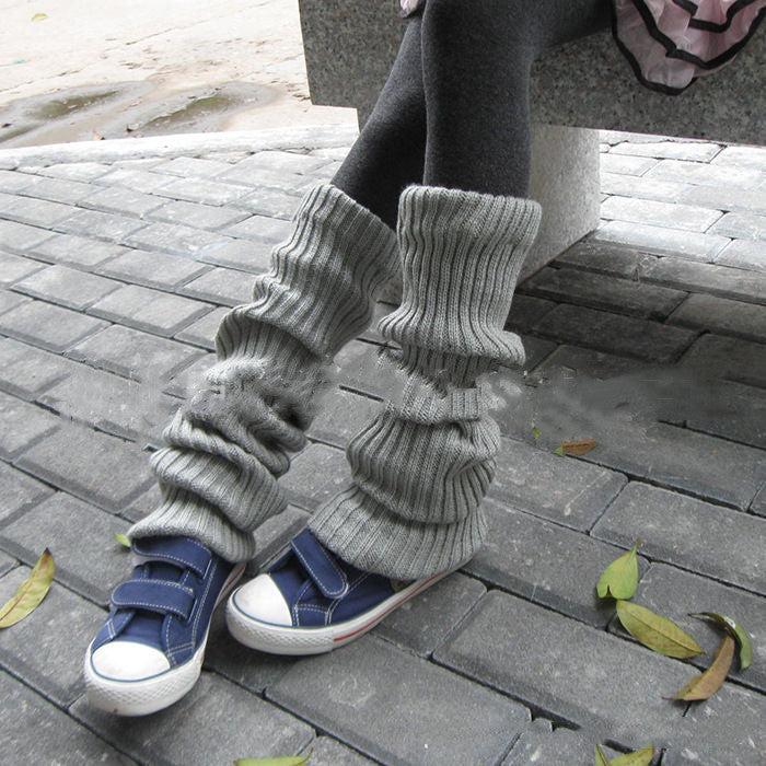 Grey knitted leg warmers worn over dark leggings and blue sneakers on a paved walkway.