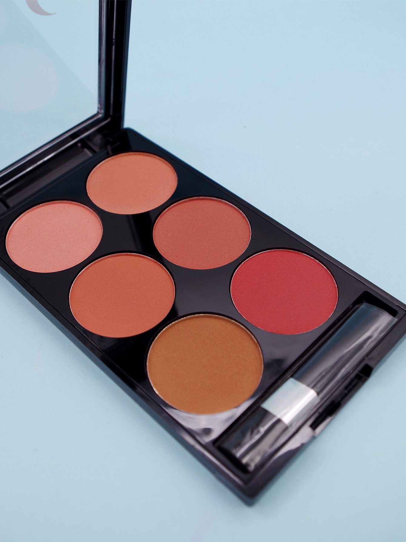 Six-color blush palette in a black case with an applicator brush, featuring various shades of pink, red, and bronze.