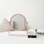 Pink circular tabletop makeup mirror with an adjustable metal frame on a white table next to cosmetics.